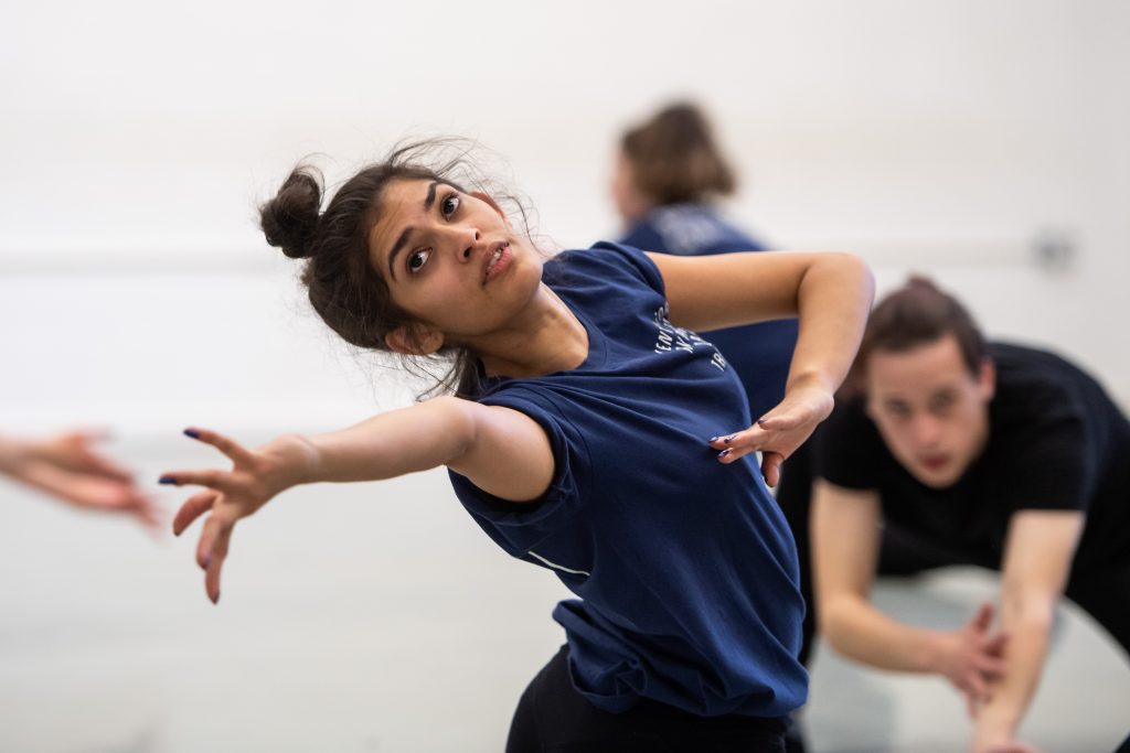 A dancer reaches backwards with one hand, their fingers are splayed, their other hand is by their chest. They are following their reach with their eyes.